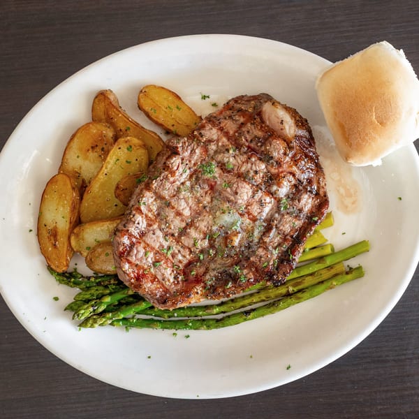 Grilled steak with roasted potato wedges, asparagus, and a dinner roll on a white plate.