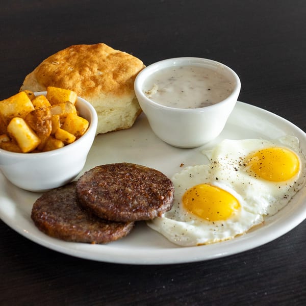 A breakfast plate with two fried eggs, two sausage patties, a biscuit, gravy, and home fries.