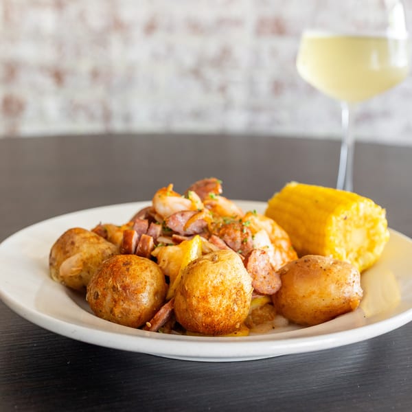 A seafood boil with shrimp, sausage, potatoes, and corn on the cob, with a glass of white wine.