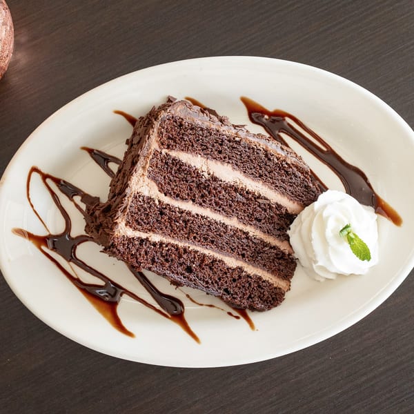 A slice of chocolate cake with chocolate frosting, chocolate drizzle, whipped cream, and a mint leaf.
